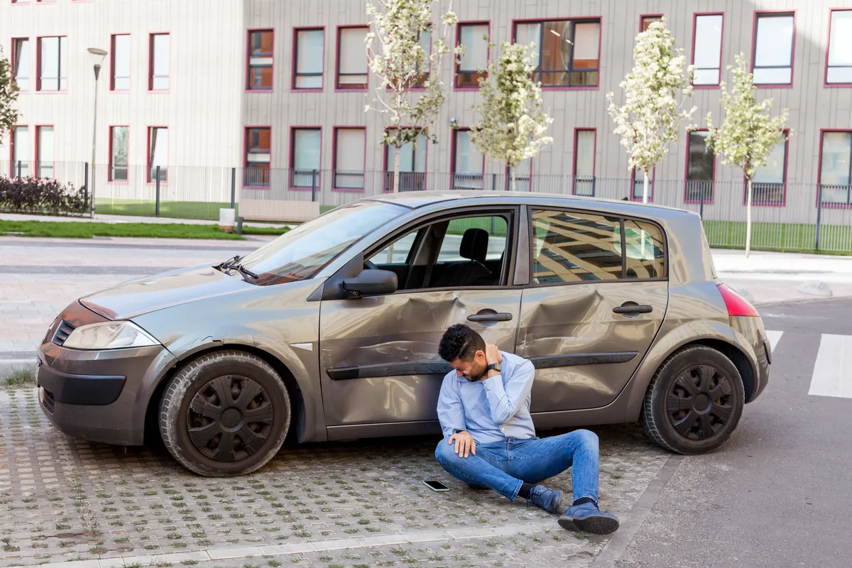 arafed man sitting on the ground next to a parked car
